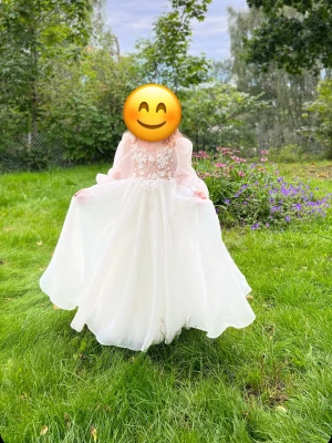 Vit långklänning med spets och volang - Drömmig vit långklänning med transparenta långärmar och vacker spets upptill. Kjolen är luftig och svepande med volangdetaljer. Överdelen har blommiga broderier och en lätt v-ringning, samt öppen rygg med dragkedja. Perfekt för speciella tillfällen.