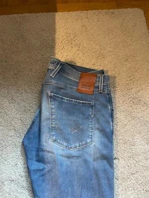 Replay Hyperflex jeans - Säljer ett par Replay Hyperflex jeans i ljusblå tvätt med slitningar och distressed detaljer på benen. Klassisk femficksmodell med skinnpatch bak i midjan. Jeansen har smal passform och är tillverkade i stretchigt jeansmaterial för extra komfort.