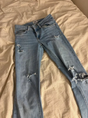 Ljusblå bootcut jeans från 157 - Ljusblå jeans från 157 med bootcut-modell och låg midja. Jeansen har slitna detaljer och hål på benen för en trendig, avslappnad look. Klassisk femficksdesign och råa kanter vid benslut. Perfekta för dig som gillar en chill och edgy stil.