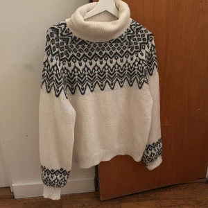 Vit polotröja med mönster - Säljer en vit polotröja med svart geometriskt mönster över axlar och ärmslut. Tröjan har en bred, ribbad polokrage och är långärmad med mjukt, stickat material. Perfekt för kyliga dagar och har en mysig, oversized känsla.