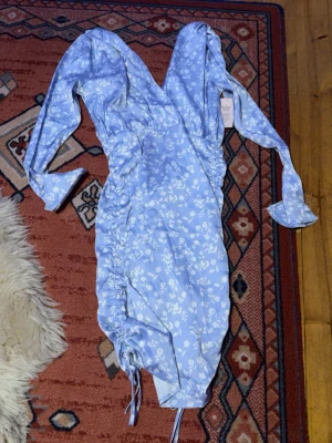  sortblå blommig kortklänning från Nelly - Supersöt ljusblå kortklänning från Nelly med vita blommor, v-ringning och långa ärmar. Klänningen har snörning i sidorna och en figurnära passform som framhäver midjan. Dragkedja på ryggen. Perfekt för sommarens alla tillfällen! Aldrig använd, endast testad. Säljes då det inte är min stil och var för stor för mig. Materialet är lite mer åt silkes hållet men är inte säker. 