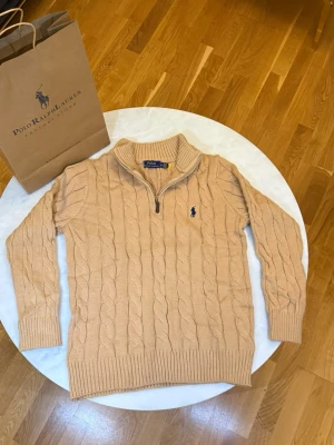 Beige stickad tröja Polo Ralph Lauren - Säljer en beige stickad tröja från Polo Ralph Lauren med klassiskt kabelmönster och half zip-dragkedja vid halsen. Tröjan har långärmad design, ribbade muddar och en broderad marinblå logga på bröstet. Perfekt för dig som gillar stilren och tidlös look.
