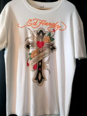 Vit Ed Hardy t-shirt med tryck - Säljer en vit t-shirt från Ed Hardy med stort färgglatt tryck på framsidan med kors, hjärta och blommor. T-shirten har orange Ed Hardy-logga både fram och bak samt detaljerad design. Klassisk rund hals och korta ärmar. Materialet är mjuk bomull. XL