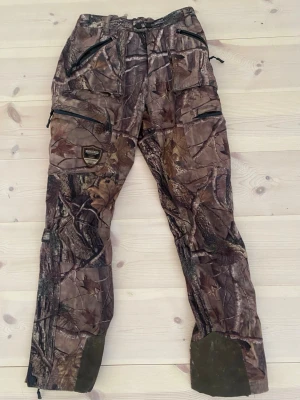 Camo cargopants från Brokared strl 36 - Säljer ett par Jaktbyxor från Brokared i camomönster. Byxorna har flera praktiska fickor med dragkedja, förstärkta benslut och ventilation. Perfekta för uteliv och äventyr. Materialet är tåligt syntet med meshfoder.