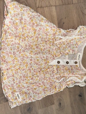 Blommig kortklänning från Newbie Newbie flerfärgade klänning, storlek 74 - Supersöt kortklänning från Newbie i storlek 74. Klänningen är vit med små blommor i rosa och gult, har volangdetaljer vid axlarna och knappar framtill. Perfekt för vår och sommar, tillverkad i mjuk bomull. Söt klänning med blommigt mönster och volangdetaljer. Klänningen har knappar framtill.