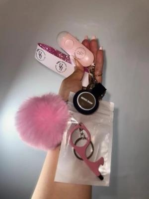 Försvarsknippa - Säljer ett nyckelsringkit med vit och rosaglittrig rem, dekorerat med en stor fluffig rosa boll, AirTag, 3-i-1 med kapsylöppnare, dörröppnare och smartphone touch och sos alarm 130 db med inbyggd lampa. Perfekt för att hålla koll på nycklar och känna dig säker. 