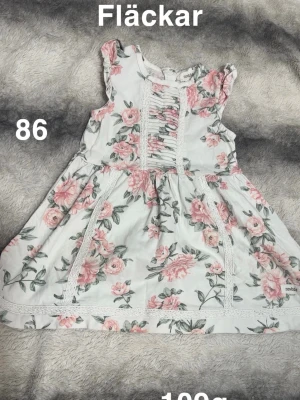 Blommig kortklänning från Newbie - Supersöt kortklänning från Newbie med blommigt mönster i rosa och grönt på vit botten. Klänningen har volangärmar, spetsdetaljer och rynk framtill. Perfekt för sommarens alla tillfällen!🌸 Fläckar storlek 86