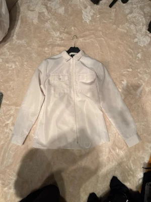 Vit overshirt från boohooMAN - Säljer en stilren vit overshirt från boohooMAN i storlek M. Jackan har två bröstfickor med knappar, dragkedja framtill och klassisk krage. Materialet är lätt och känns fräscht, perfekt för lager-på-lager. Passar dig som gillar enkel och clean stil.