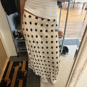 Prickig långkjol i satin - Snygg långkjol med svart prickigt mönster på ljus beige botten. Kjolen har en rak och flowy passform som ger en cool och avslappnad vibe. Materialet är glansigt satin som känns mjukt mot huden och ger en lyxig känsla. Perfekt för dig som gillar statement-plagg.