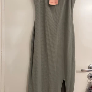 Ribbad grön långklänning med slits - Snygg ribbad långklänning i grönt med en stilren slits framtill. Klänningen har en tight passform och är perfekt för dig som gillar minimalistisk stil. Materialet är stretchigt och följsamt, vilket gör den både bekväm och trendig.