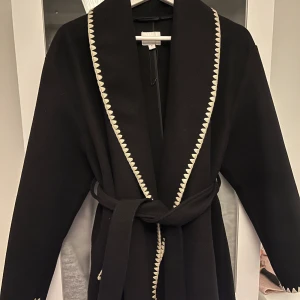 Svart kappa med mönstrad kant Kappahl - Svart kappa från Kappahl i storlek S med bred krage och dekorativ vit sicksackkant längs krage, fickor och ärmslut. Kappan har knytbälte i midjan och två fickor framtill. Perfekt för dig som gillar stilrena detaljer och vill ha en snygg look.