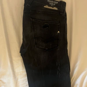 Svarta Replay jeans med slitningar - Säljer ett par svarta jeans från Replay med coola slitningar och distressed detaljer både fram och bak. Modellen heter Broken Edge och har klassisk femficksdesign. Jeansen har en snygg mörk tvätt och är perfekta för en avslappnad streetstil.