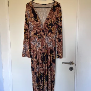 Mönstrad jumpsuit från NLY One - Säljer en maxad jumpsuit från NLY One i storlek XS med v-ringad omlottfront och långa ärmar. Klänningen har ett unikt mönster med leopard och barockinspirerade detaljer i brunt, beige och svart. Materialet är mjukt och stretchigt, perfekt för dig som gillar att sticka ut.