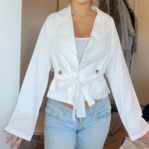 Vit tunn jacka med knytband - Snygg vit tunn jacka med bred krage, knytband i midjan och dekorativa knappar framtill. Jackan har långa ärmar och en stilren look som passar perfekt till jeans eller kjol. Materialet känns lätt och fräscht, perfekt för vår och sommar.