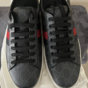 Gucci Ace skor svarta  - Gucci Ace sneakers i svart skinn med GG-monogram, röda och blå ränder på sidan samt olika färg på hälarna – en röd och en blå i metallic. Klassisk vit sula med unika detaljer undertill. Svarta skosnören och rund tå. Perfekta för dig som vill sticka ut med lyxig. Har box och Dust bag skorna e helt nya. Skriv vid frågor kan diskutera priset 