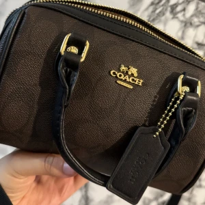 Coach svart handväska i skinn - Snygg och stilren svart handväska från Coach i äkta skinn med guldfärgade detaljer och dragkedja. Väskan har två handtag och klassisk Coach-tag. Perfekt storlek för det viktigaste och med lyxig känsla.