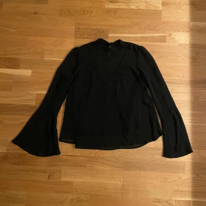 Svart blus med utvsvängda ärmar - Snygg svart blus med långärmade utsvängda ärmar och liten ståkrage. Blusen har en lätt och flowy passform, perfekt för att lyfta din outfit med en stilren touch.