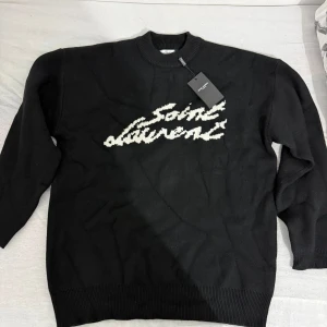 Svart stickad Saint Laurent tröja - Svart stickad tröja från Saint Laurent med vit broderad logga framtill. Klassisk rund halsringning och ribbade muddar vid ärmslut och nederkant. Perfekt för dig som vill ha en stilren och exklusiv look.