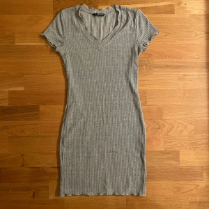 Grå ribbad kortklänning  - Superenkel och snygg grå ribbad klänning med v-ringning och korta ärmar. Den är figurnära och har en mjuk, stretchig känsla som gör den bekväm att bära. Perfekt för dig som gillar minimalistisk stil och vill ha något lättmatchat.