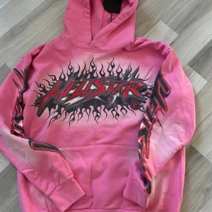 Rosa Hellstar hoodie med flames - Säljer en rosa hoodie från Hellstar med fet flames-grafik och logga på bröstet och armarna. Baksidan har en stor svart siluett med stjärna och flames. Hoodien har huva och är tillverkad i mjuk bomull. Perfekt statement-plagg för dig som gillar streetwear.