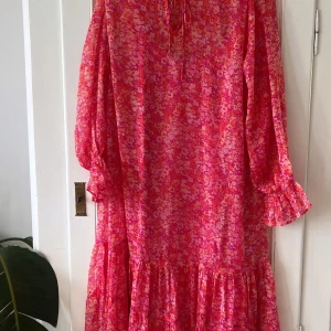Rosa blommig långklänning med volang - Säljer en färgstark rosa långklänning med blommigt mönster och volangdetaljer längst ner. Klänningen har långa ärmar med rynkade muddar och knytning vid halsen. Perfekt för dig som gillar boho-vibe och vill sticka ut med mycket färg.