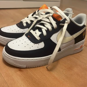 Nike Air Force 1 sneakers blå/vit - Snygga Nike Air Force 1 sneakers i blått och vitt med krämfärgade detaljer och orangea accenter. Skorna har klassisk låg siluett, snörning och en cool sol-emoji på hälen. Tillverkade i skinn och syntet, med perforerad tå och vit sula för extra stilpoäng.
