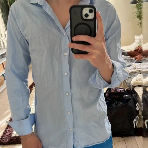 Ljusblå skjorta från The Shirt Factory - Snygg ljusblå skjorta från The Shirt Factory i slim fit-modell. Skjortan är tillverkad i bomull och har klassisk krage, långa ärmar och vita knappar framtill. Perfekt för dig som gillar stilrena och enkla plagg med modern passform.