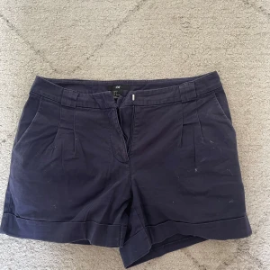Mörkblå shorts från H&M, stl 36 - Snygga mörkblå shorts från H&M med klassisk design och uppvikta benslut. De har fickor fram och bak, bälteshällor och stängs med dragkedja. Materialet är mjukt och bekvämt bomullstyg.