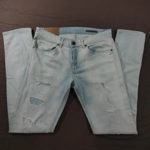 Ljusblå Dondup  Jeans med slitningar - Säljer ett par ljusblå jeans från Dondup med slitna detaljer och hål på benen. Jeansen har fem fickor, knappgylf och klassisk, slim passform. Perfekta för en snygg och trendig look.