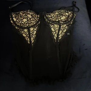 Svart spetskorsett med dragkedja - Säljer en svart korsett med tunna axelband och vacker spetsdetalj upptill. Toppen har meshpartier och dekorativa volanger nedtill. Baksidan har en praktisk dragkedja för enkel på- och avtagning. Perfekt för dig som vill ha en edgy och snygg look.