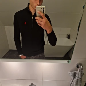 Svart stickad half zip från Polo Ralph Lauren - Svart stickad tröja från Polo Ralph Lauren med röd broderad logga på bröstet. Tröjan har half zip-dragkedja, ståkrage och lång ärm. Perfekt för lager-på-lager och snygg till jeans. Klassisk och stilren design.