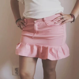 Rosa kortkjol med volangkant - Supersöt rosa kortkjol med volang längst ner och klassisk jeansmodell med fickor och knapp framtill. Kjolen har en ljus rosa ton och är perfekt för dig som gillar en feminin och lekfull stil. Passformen är normal och materialet känns som mjuk bomull.