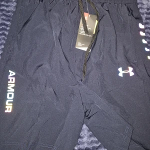 Svarta träningsshorts från Under Armour - Snygga svarta träningsshorts från Under Armour med regnbågsfärgad logga och text på benet. Shortsen har elastisk midja med snörning och är gjorda i ett lätt och svalt polyestermaterial. Perfekta för gymmet eller löprundan.