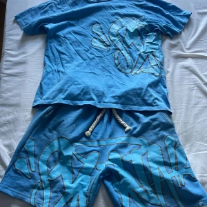 Syna world suit  - Säljer en blå syna world suit med stort grafiskt tryck i ljusblått och vitt på både fram- och baksidan. T-shirten har rund hals, korta ärmar och en avslappnad passform. Materialet känns mjukt och skönt, perfekt för varma dagar.