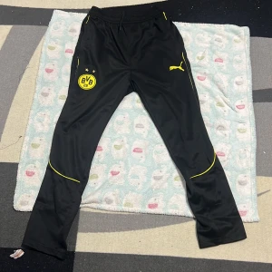 Svarta BVB träningsbyxor från Puma S  - Svarta träningsbyxor från Puma med Borussia Dortmunds BVB-logga och gula detaljer. Byxorna har elastisk midja, smal passform och är tillverkade i 100% polyester. Perfekta för fotbollsträning eller chill. Har inte blivit använt en enda gång men saknar tagg 