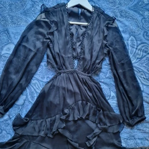 Svart klänning med volanger från Asos - Supersnygg svart klänning med långa transparenta ärmar, djup v-ringning och cut out del med dekorativa volanger. Använd 1 gång.