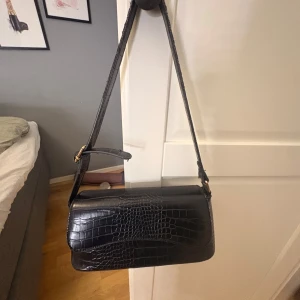 Svart axelväska i croco-skinn - Snygg svart axelväska med croco-mönster och justerbar rem. Väskan har ett stilrent, rektangulärt format och insida med dragkedja för extra säkerhet. Perfekt storlek för mobil, plånbok och andra essentials. Materialet är skinn med en cool textur som ger en lyxig vibe.