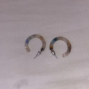 Färgglada hoops örhängen - Säljer ett par coola hoops örhängen i akryl med mixade färger som beige, blått och brunt. De har en halvcirkelform och metallfäste. Perfekta för att ge din look lite extra edge!