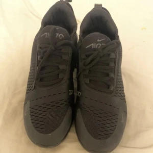 Nike Air Max 270 svarta sneakers - Säljer ett par svarta Nike Air Max 270 sneakers med snygg, sportig design. Skorna har mesh-överdel för bra andningsförmåga, synlig Air Max-dämpning i hälen och klassisk snörning. Perfekta för dig som gillar streetwear och vill ha bekväma skor med modern look.