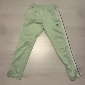Mintgröna träningsbyxor från Lacoste - Snygga mintgröna träningsbyxor från Lacoste med vita sidoränder och dragkedjor vid fickorna. Byxorna har elastisk midja med snörning och är tillverkade i ett glansigt syntetmaterial. Perfekta för en sportig look.