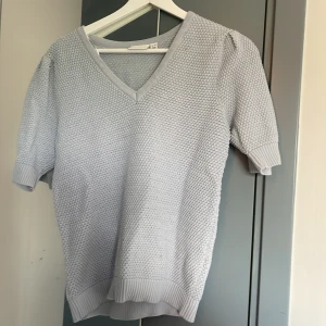 Ljusgrå stickad v-ringad tröja - Säljer en ljusgrå stickad tröja med v-ringning och korta ärmar. Tröjan har en grovstickad struktur och ribbade muddar vid ärmslut och nederkant. Perfekt för lager-på-lager och enkel att matcha med jeans eller kjol.