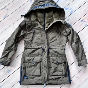 Olivgrön parkajacka Sail Racing M - Parkajacka från Sail Racing i olivgrönt med stora fickor, justerbar midja med snörning och dragkedja framtill. Jackan har huva med logga, flera dragkedjeförsedda fickor och fodrad insida för extra värme. Perfekt för kalla dagar och snygg streetstil. Nu pris ca 4000 kr. Väldigt fint skick. 