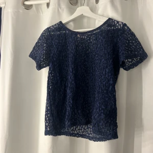 Mörkblå spetsblus från Modström - Säljer en mörkblå blus från Modström i genombruten spets med korta ärmar. Blusen har rund halsringning med röd kant och ett snyggt mönster i spetsen som ger en cool look. Perfekt att styla med jeans eller kjol för en trendig outfit.
