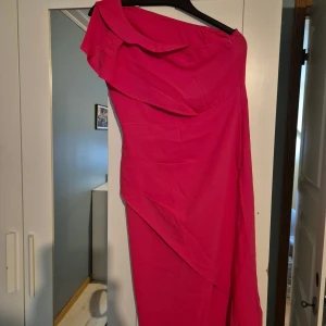 Chockrosa oneshoulder klänning - Säljer en snygg chockrosa klänning med oneshoulder-design och volangdetalj upptill. Klänningen har en asymmetrisk skärning och är figurnära, perfekt för dig som vill sticka ut. Materialet känns mjukt och följsamt mot kroppen.