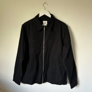 Svart overshirt från Kin i bomull - Stilren svart overshirt från Kin i 100% bomull. Jackan har raka linjer, två bröstfickor och hel dragkedja framtill. Klassisk krage och långa ärmar med knapp vid ärmslut. Perfekt för lager-på-lager och enkel att matcha med allt. 3U2