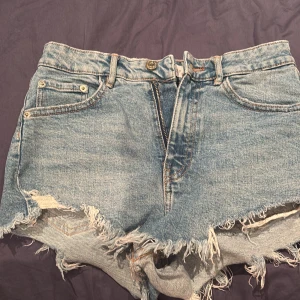 Blå jeansshorts med fransig kant från Zara - Säljer ett par blå jeansshorts från Zara i storlek 34. De har rå, fransig kant nertill och klassisk femficksmodell. Shortsen har dragkedja och knapp framtill samt bälteshällor. Perfekta för en avslappnad sommarstil.