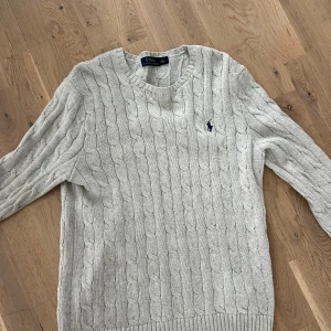 Ljusgrå kabelstickad tröja Polo Ralph Lauren - Kabelstickad tröja från Polo Ralph Lauren i ljusgrått med klassisk rund halsringning och broderad marinblå logga på bröstet. Mjuk och skön, perfekt för lager-på-lager. Långärmad och ribbade muddar för extra stilpoäng. Den är lite större i storleken! 😊