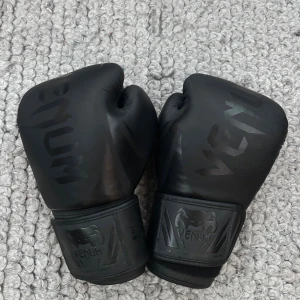 Svarta Thaiboxningshandskar från Venum - Säljer ett par svarta Thaiboxningshandskar från Venum med stilren matt finish och diskret logga på ovansidan och vid handleden. Handskarna har en klassisk form och bred kardborrestängning för extra stöd.