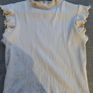 Ribbad ärmlös topp med volang från Zara - Supersöt ribbad topp från Zara i ljusbeige med volangdetaljer vid ärmöppningarna och en liten volangkrage. Toppen är ärmlös och har en tight passform som ger en fräsch och trendig look. Perfekt att styla med jeans eller kjol för en chic vibe.