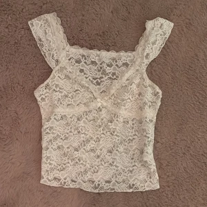 Vit spetsig linnetopp med volang - Söt vit linnetopp i spets med v-ringning och små volangdetaljer vid axelbanden. Toppen är ärmlös och har ett romantiskt mönster i spets över hela plagget. Perfekt för dig som gillar en feminin och trendig stil.